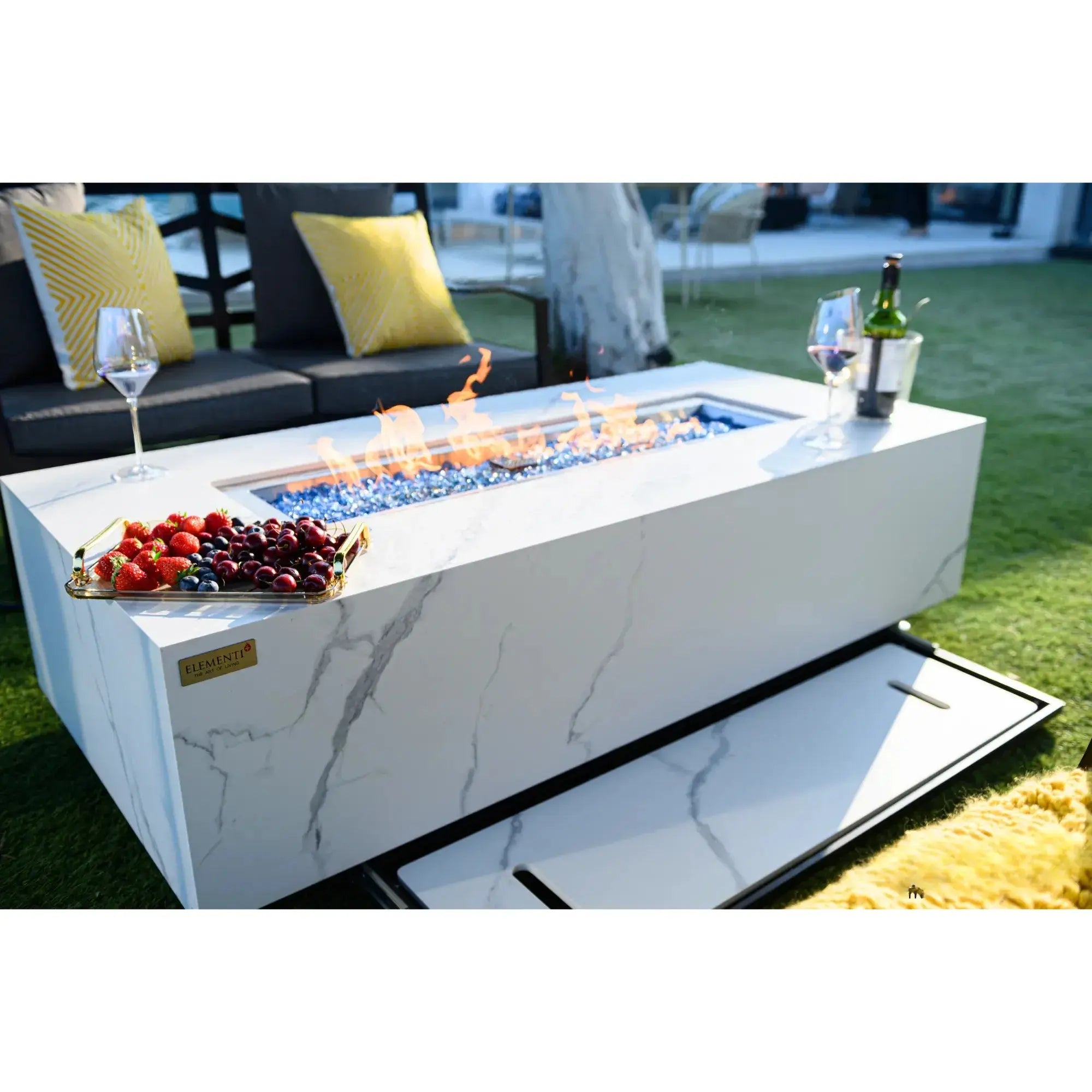 Outdoor fire pit table with flames, fruit, and drinks on a grassy area.