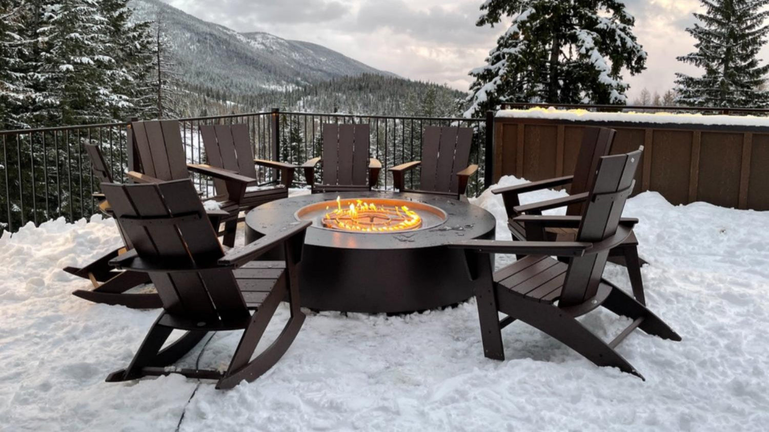 Outdoor fire pit table with chairs on a snowy deck with mountains in the background