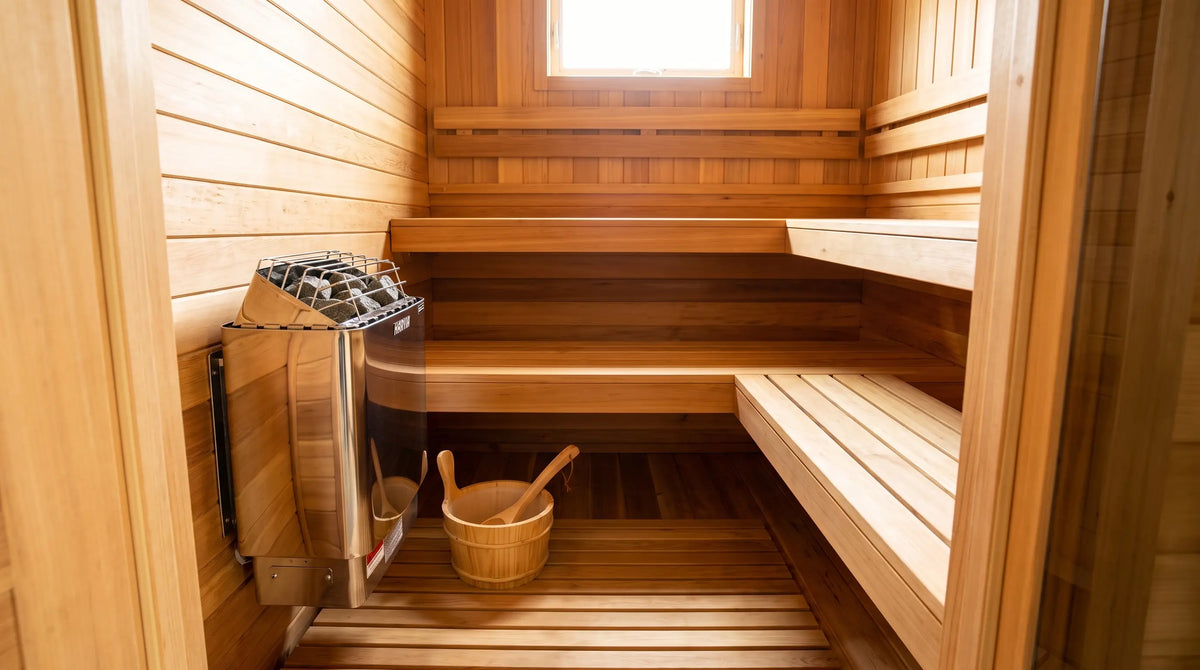 Wide view from the doorway of a compact cedar sauna with a Harvia KIP wall heater, tiered benches and a wooden bucket