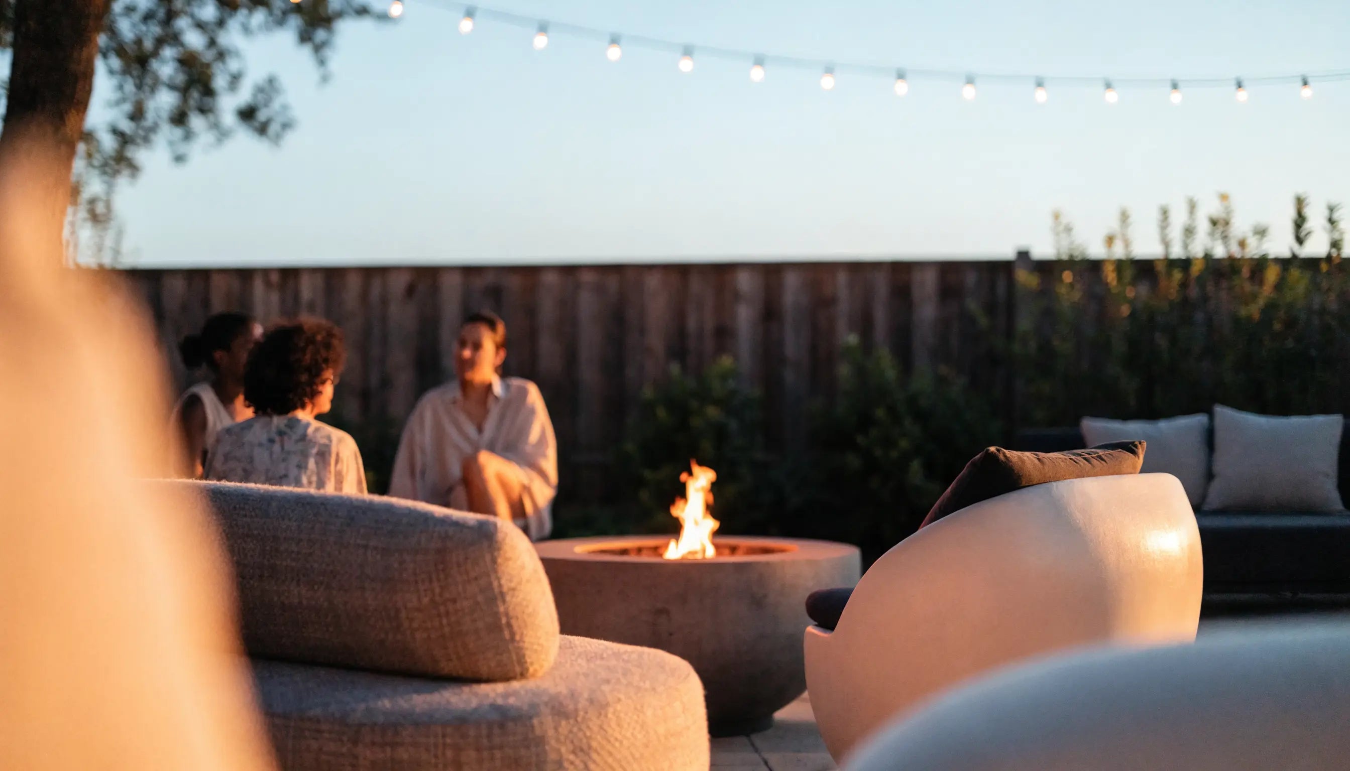 Outdoor fire pit glowing on a backyard patio at dusk with lounge furniture and string lights