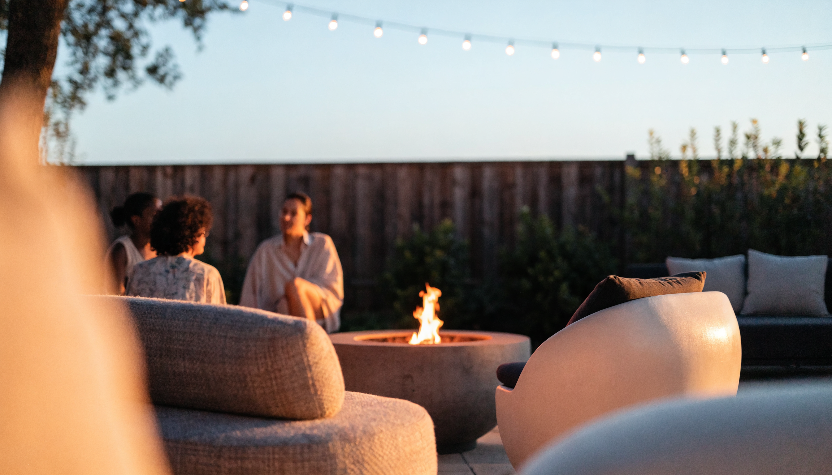 Outdoor fire pit glowing on a backyard patio at dusk with lounge furniture and string lights